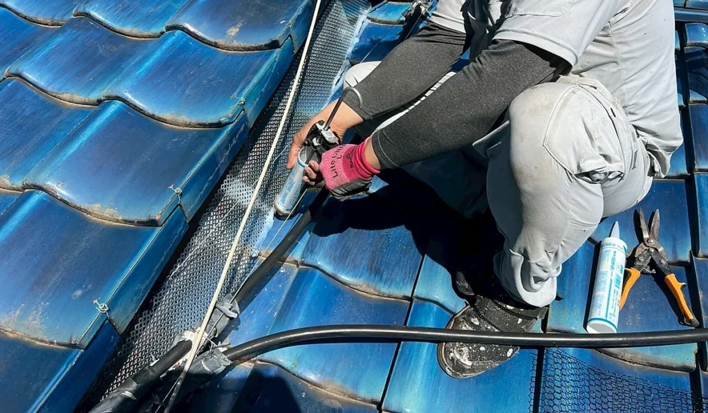 青い瓦屋根で、穴あきの金属板の縁にコーキングを打って塞いでいる職人の手元