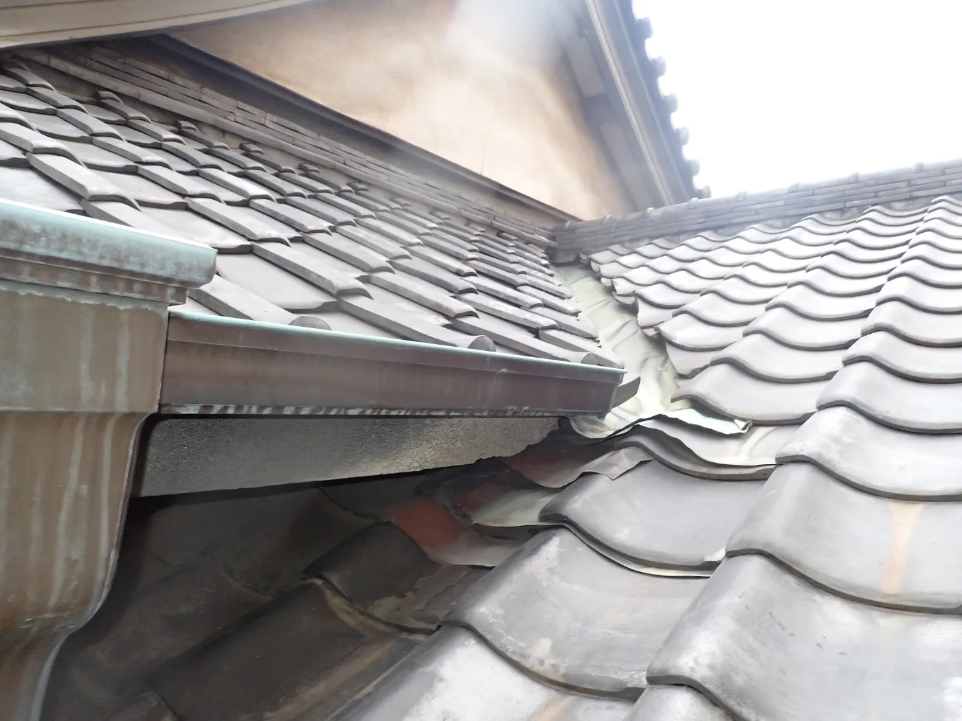 軒先の雨樋の奥で、上下の瓦屋根と外壁が重なり、その下に暗いすき間が見える屋根の端
