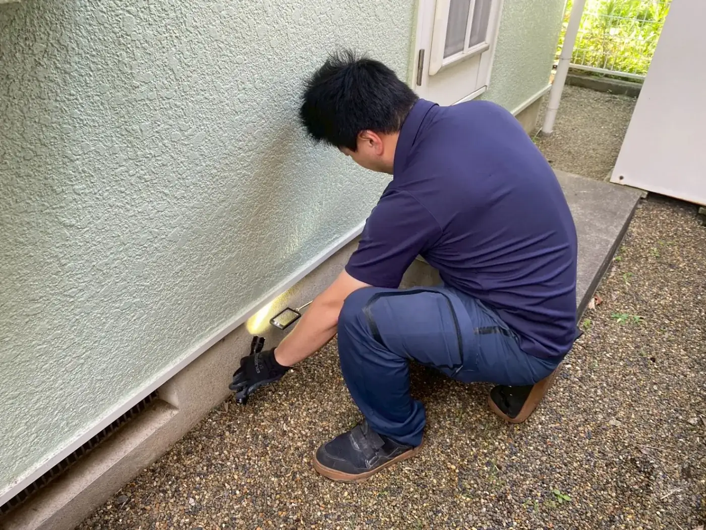 外壁の基礎の際にしゃがみ、ライトを当てて床下通気口のまわりを調べる作業員