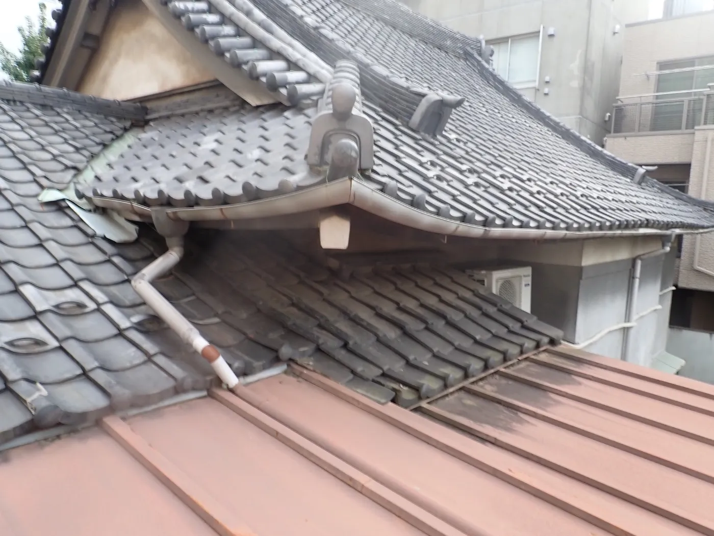 二つの瓦屋根が重なる谷と、手前の金属屋根へ下りる雨どいを上から見た建物