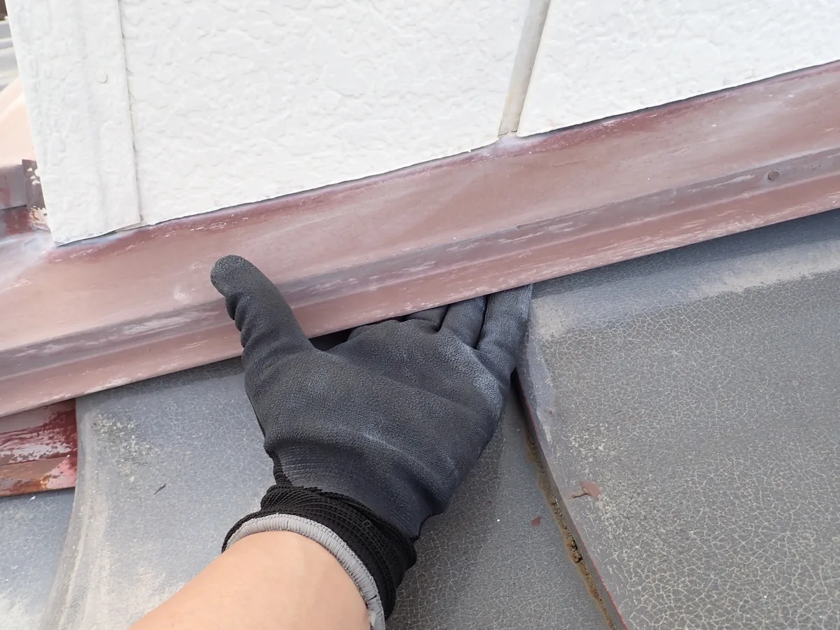 灰色の屋根材と外壁下の板金の間にできた隙間へ、手袋の手を差し入れて示している調査の場面