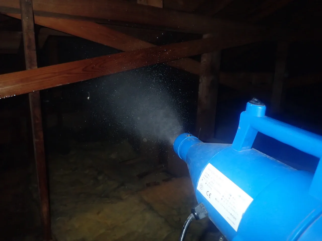 屋根裏の梁の下で、青い噴霧器から薬剤を霧にして吹き付ける作業