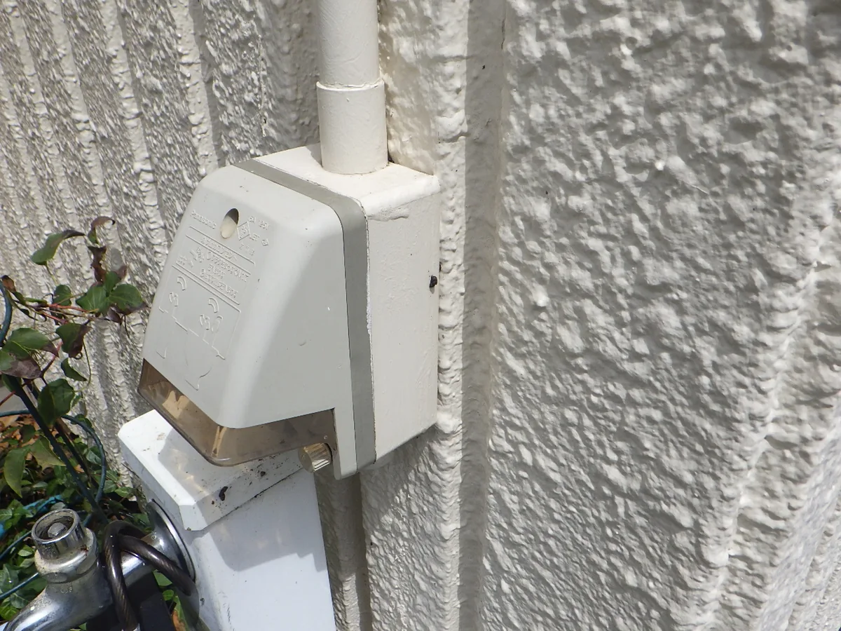 吹き付け仕上げの外壁に付いた白い電気ボックスと、上へ延びる配管。ボックスの側面に小さな黒い穴が見える