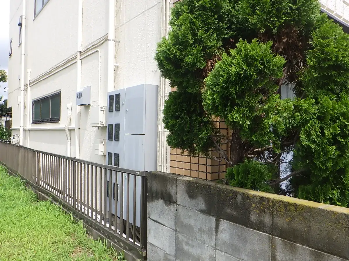 外壁のすぐそばまで植木が茂り、ブロック塀とフェンスに囲まれた建物の周辺