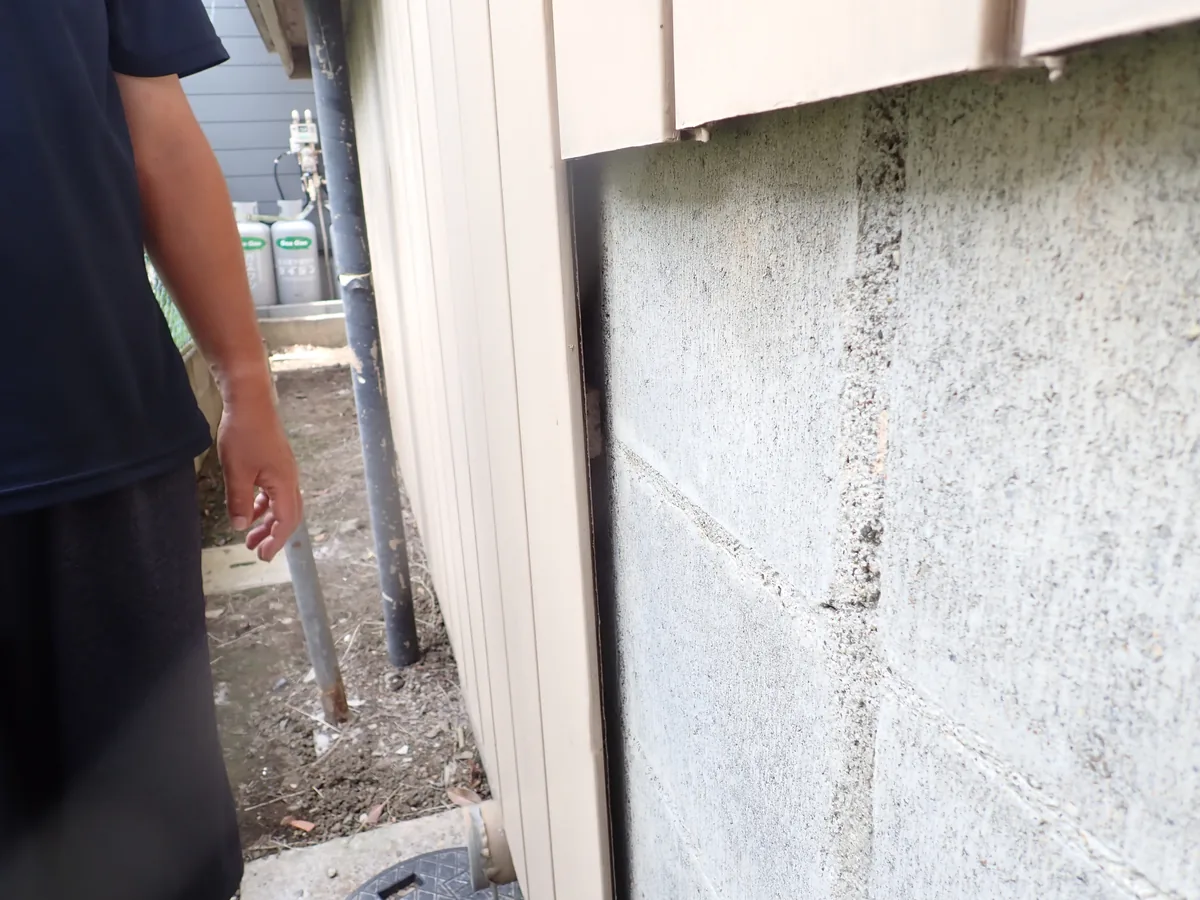 外回りの点検中に見つけた、ブロック塀と建物の外板の間の細いすき間と、脇に立つ作業員