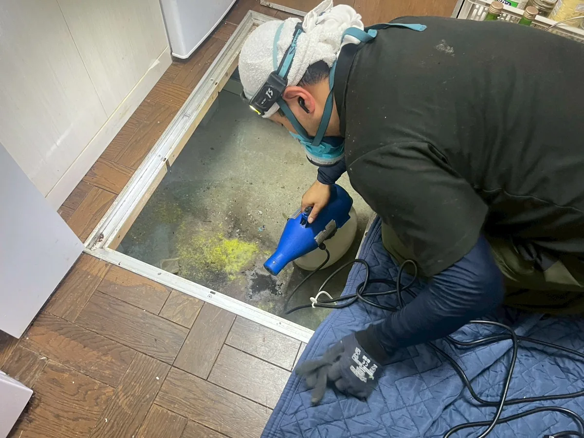 防護具を着けた作業員が床下点検口から噴霧器を入れ、床下の消毒を行っている様子