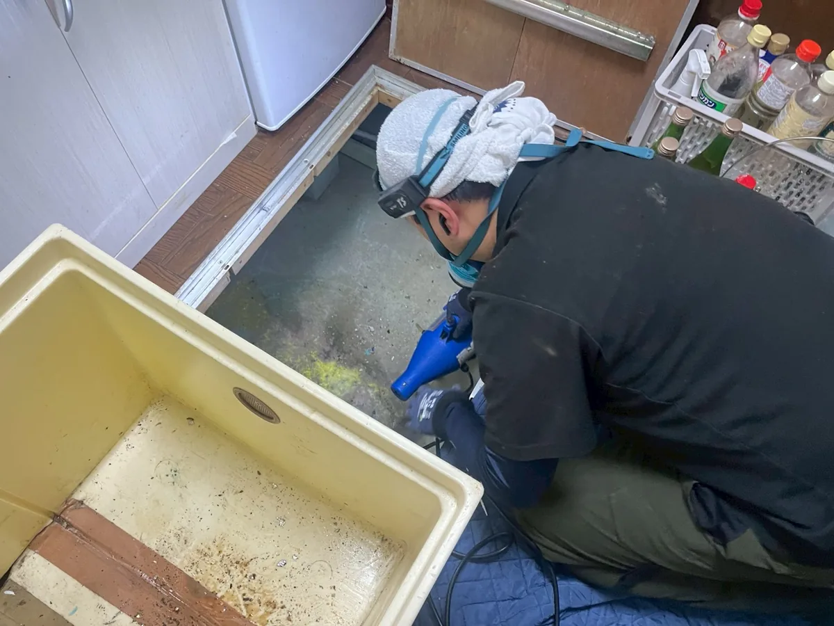 台所の床下点検口で、作業員が青い噴霧器の先を床下へ向けて薬剤をまいているところ